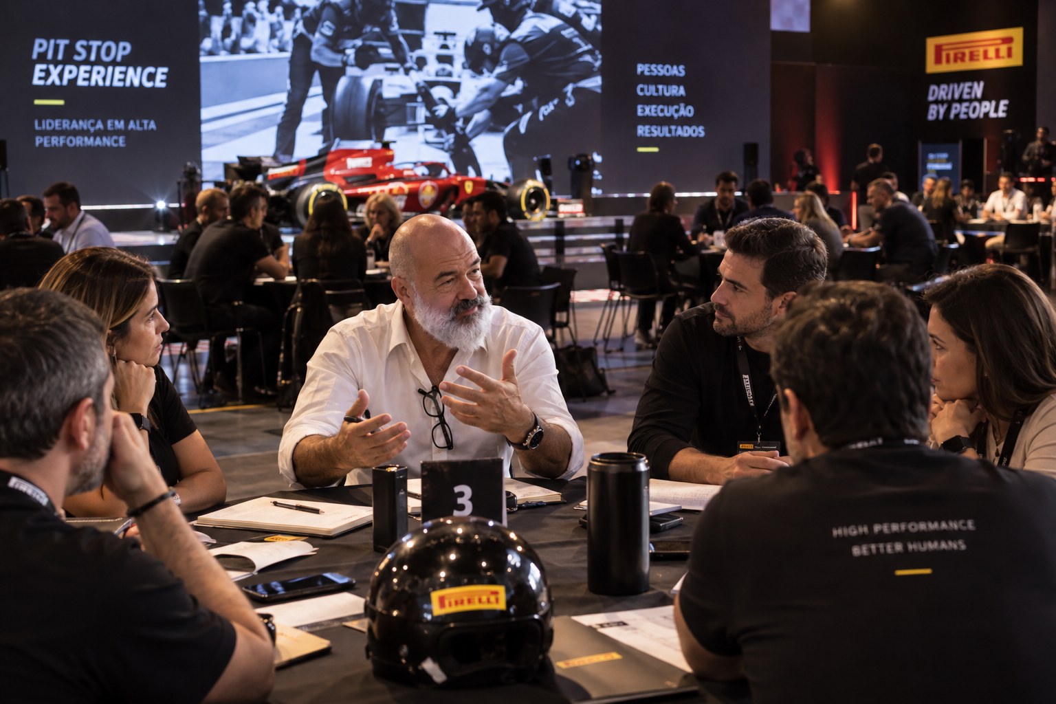Cadu Lemos conduz o debriefing com uma equipe à mesa, telão ao fundo com os dizeres Pit Stop Experience — Liderança em alta performance